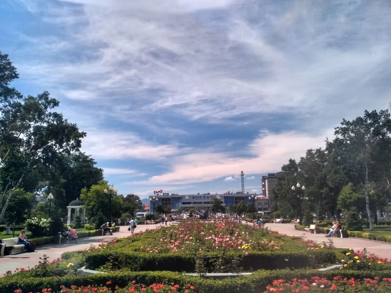 За клумбой мемориал жертвам землетрясения в Нефтегорске, на заднем плане здание железнодорожного вокзала